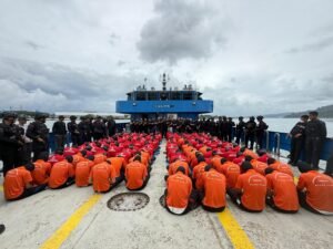 Narapidana berbaju oranye dan merah duduk berbaris di geladak kapal penyeberangan ke Pulau Nusakambangan dengan penjagaan petugas bersenjata. 
