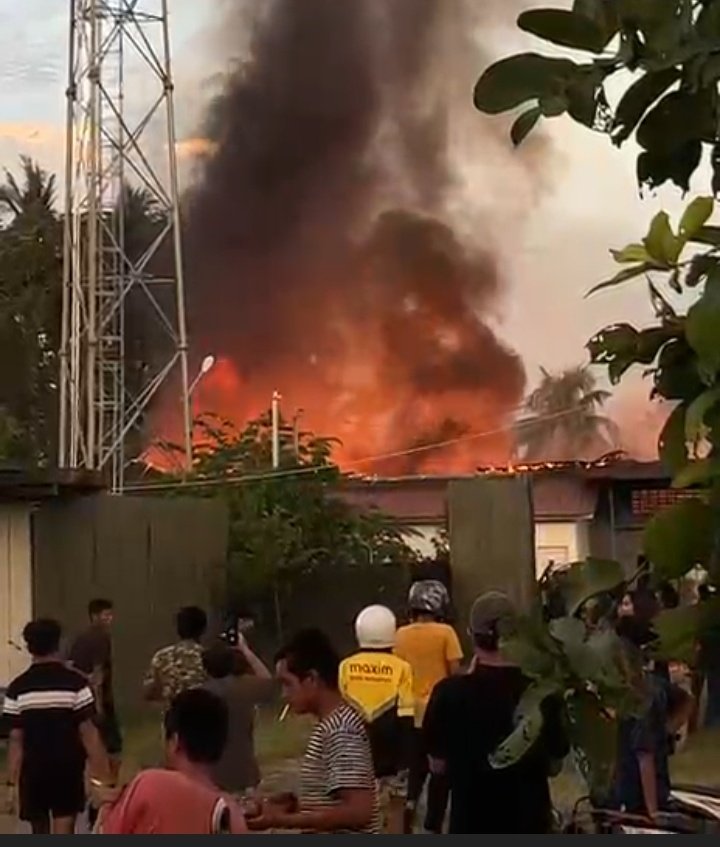 Kebakaran hebat melanda pemukiman warga di Rawang, Padang Selatan, memperlihatkan kobaran api besar dan kepulan asap hitam membumbung tinggi di dekat tower telekomunikasi sementara warga berkumpul menyaksikan kejadian.