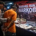 Polisi mengamankan tersangka narkoba di depan meja berisi barang bukti sabu, uang tunai, dan timbangan dengan latar tulisan “Barang Bukti Narkoba”.
